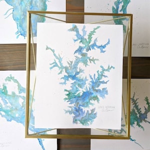 May include: Watercolor painting of Lake Norman in shades of blue and green, framed in a gold metal frame. The artwork is on white paper with the lake's outline. The text "Lake Norman" is visible. Other similar paintings are in the background.
