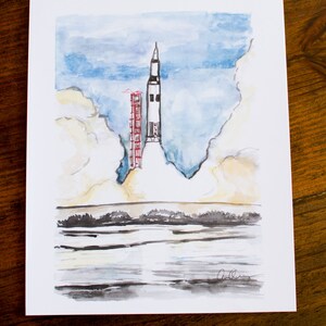 May include: A watercolor painting of a rocket launching into a cloudy sky. The rocket is white with red accents and is surrounded by white clouds. The sky is blue with white clouds. The painting is signed in the bottom right corner.