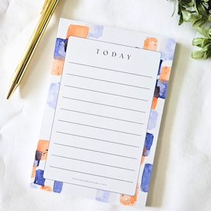 May include: A white notepad with the word "TODAY" at the top, featuring lined pages for notes. The notepad is decorated with watercolor-style orange and blue squares. A gold pen rests on the side.