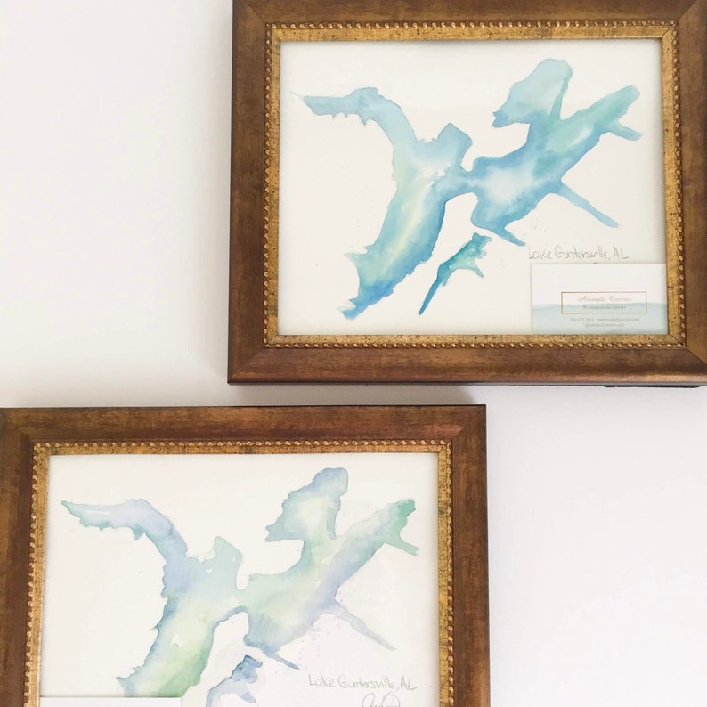 May include: Two framed watercolor paintings of Lake Guntersville, Alabama. The paintings are in shades of blue and green, and are framed in brown wood with gold accents. The text "Lake Guntersville, AL" is written in cursive at the bottom of each painting.