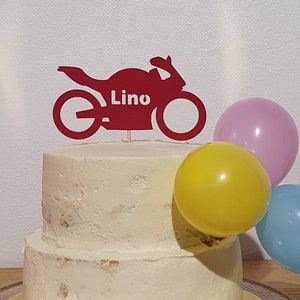 Puede incluir: Un pastel blanco de dos pisos con un adorno de motocicleta roja que dice "Lino". El pastel está decorado con tres globos: uno rosa, uno amarillo y uno azul.