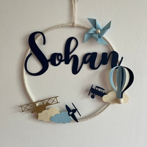 Puede incluir: Un círculo de cuerda blanca con el nombre "Sohan" en letras negras. El círculo está decorado con aviones de papel azul y blanco, un globo aerostático de papel azul y blanco, un molino de viento de papel azul y blanco y nubes blancas.
