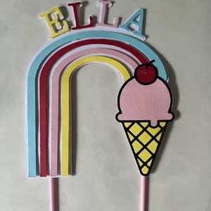 May include: A cake topper with the name "ELLA" in yellow, red, and blue letters above a rainbow design. A pink ice cream cone with a cherry on top is also included. The topper is supported by two pink sticks.