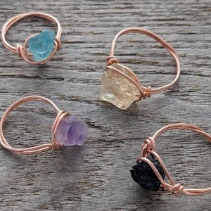 May include: Four wire-wrapped rings with different colored stones. The rings are made of rose gold wire and feature a light blue, yellow, purple, and black stone.