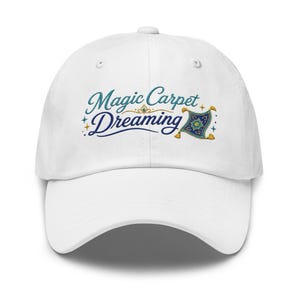 Puede incluir: Una gorra de béisbol blanca con un diseño bordado en la parte delantera. El texto Magic Carpet Dreaming aparece en letra cursiva de color verde azulado y azul oscuro. Incluye una pequeña ilustración detallada de una alfombra mágica con borlas.