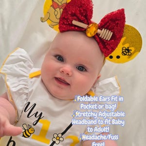 May include: A baby wearing a headband with Winnie the Pooh ears and a red sequined bow. The baby is also wearing a white onesie with yellow accents and the text "My 1st Birthday". The image includes text that says "Foldable Ears Fit in Pocket or bag! Stretchy Adjustable Headband to fit Baby to Adult! Headache/Fuss Free!"
