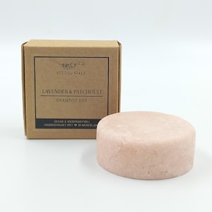 Op de afbeelding: Een lichtroze, ronde shampoo bar naast een bruine kartonnen doos. De doos heeft de tekst "STUDIO NI LE" en "LAVENDER & PATCHOULI SHAMPOO BAR". De doos vermeldt ook "VEGAN & DIERPROEFVRIJ HANDGEMAAKT MET IN NEDERLAN".