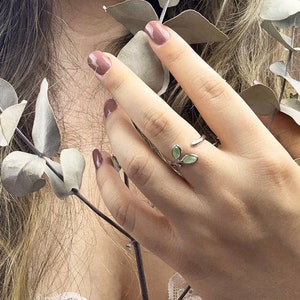 May include: A silver ring featuring two green leaves, worn on a finger. The ring is set against a backdrop of a person's hand and a sprig of eucalyptus leaves.