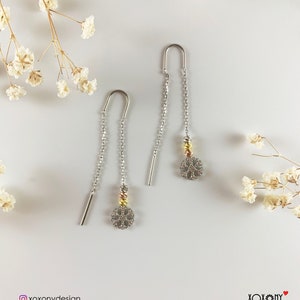 May include: A pair of silver dangle earrings with a delicate, circular design. The earrings feature a small chain with a gold and silver bead at the bottom. The earrings are photographed against a white background with dried flowers.