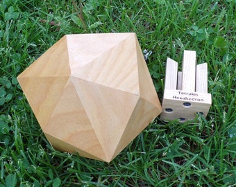 May include: Two wooden geometric shapes, a large polyhedron and a smaller cube with vertical wooden pieces, rest on green grass. The smaller cube has the text "Tetrakis Hexahedron" on it. The larger shape is a light brown color.