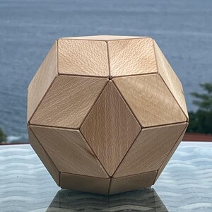 May include: A geometric polyhedral object constructed from light-colored wood with a visible grain. The shape features multiple diamond and triangular facets joined together to form a symmetrical, multi-sided solid structure with smooth edges.