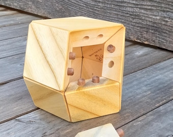 May include: A disassembled wooden puzzle cube with a light wood finish. The cube has multiple triangular faces and small wooden pegs. One piece is separated from the main structure, revealing the interior. The puzzle is set against a weathered wood background.