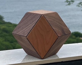 May include: A geometric polyhedral object constructed from alternating light and dark brown wood panels. The structure features a central diamond-shaped light wood face flanked by dark wood panels, creating a multi-faceted three-dimensional shape.
