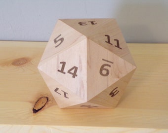 May include: A large, wooden twenty-sided die with dark brown numbers. The die is made of light-colored wood and sits on a wooden surface. The numbers are clearly visible, making it suitable for tabletop gaming.