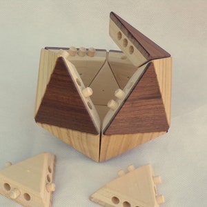 Peut inclure: Une boîte à puzzle en bois de forme géométrique. Elle est faite de bois clair et de bois plus foncé. Plusieurs pièces triangulaires sont détachées. La boîte est partiellement ouverte, révélant l'intérieur.
