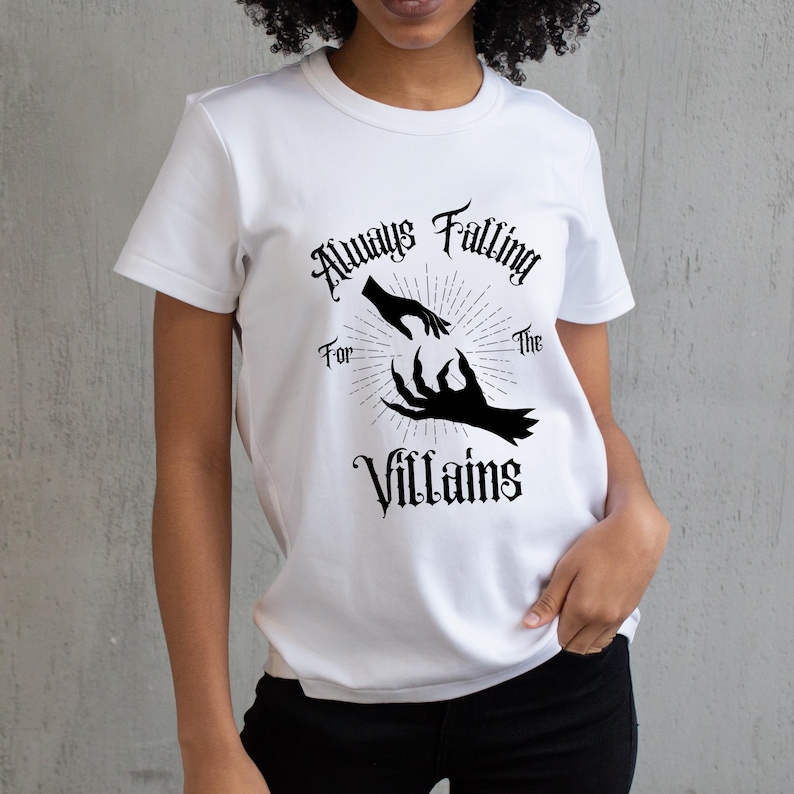 May include: White t-shirt with the text "Always Falling For The Villains" in a gothic font. The graphic features a pair of hands reaching towards each other, one human and one clawed, with a sunburst design.