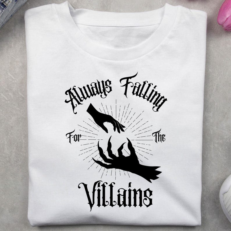 May include: White t-shirt with the text "Always Falling For The Villains" in a gothic font. The design features two hands, one reaching out and the other a clawed hand, with a sunburst design.