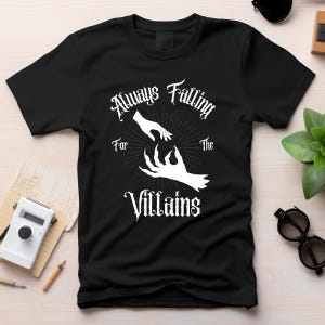 May include: Black t-shirt with a white graphic design featuring two hands reaching towards each other, with the text "Always Falling For The Villains" in a gothic font.