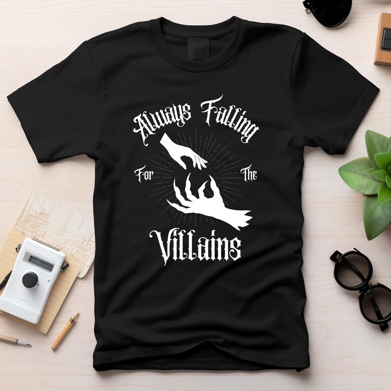 May include: Black t-shirt with a white graphic design featuring two hands reaching towards each other, with the text "Always Falling For The Villains" in a gothic font.