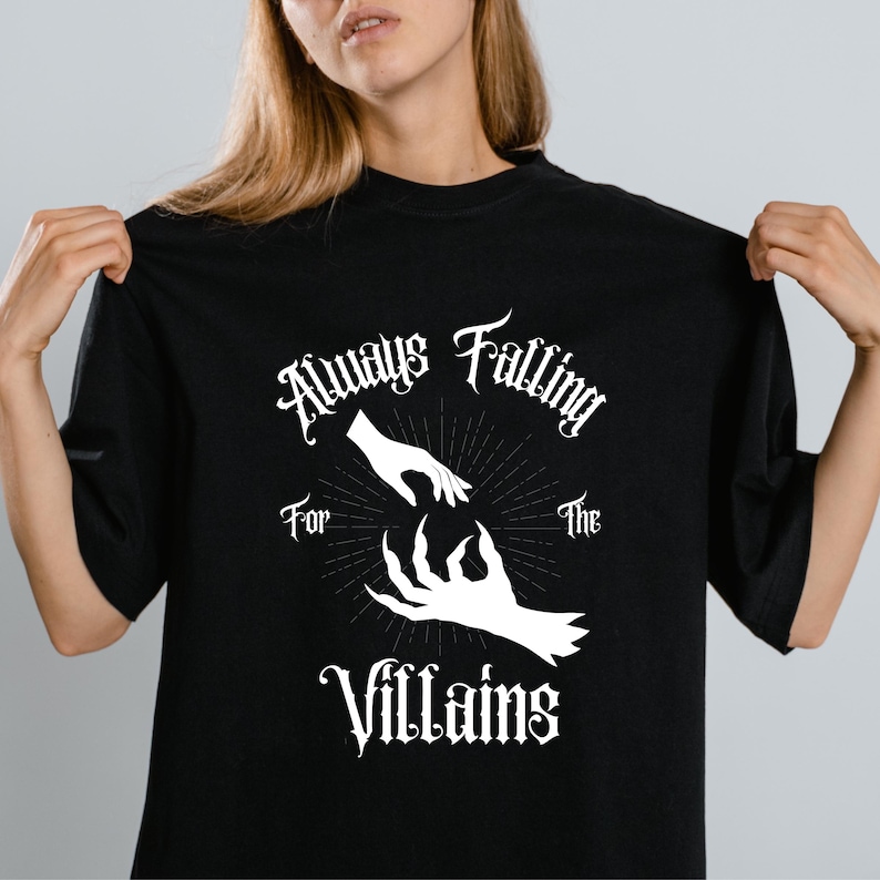 May include: Black t-shirt with the text "Always Falling for the Villains" in a gothic font. The design features a hand reaching towards a clawed hand, with a sunburst design in the center.