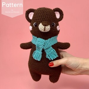 May include: A dark brown crocheted bear toy featuring a light beige muzzle, pink cheeks, and inner ears, wearing a bright turquoise scarf with fringed ends. The text Pattern by Beardie and Blondie is visible in the upper left corner.