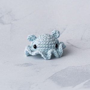 Puede incluir: Un pulpo de crochet azul claro con ojos negros. El pulpo está sentado sobre una superficie gris claro.