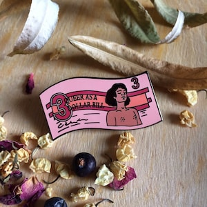 May include: Pink enamel pin shaped like a dollar bill with the text "Queer as a Dollar Bill" and a cartoon illustration of a person with short black hair and a closed-eye expression.