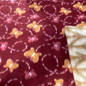 Può includere: Una coperta in pile bordeaux con un motivo ripetuto di farfalle gialle e rosa, delineate da linee tratteggiate bianche. L'angolo rivela un pile color crema con un disegno a foglie.