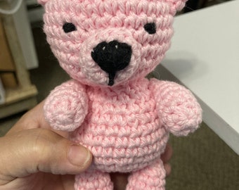 May include: A handmade pink crochet teddy bear with black eyes and a black nose. The bear has small arms, legs, and ears. The bear is approximately 6 inches tall.