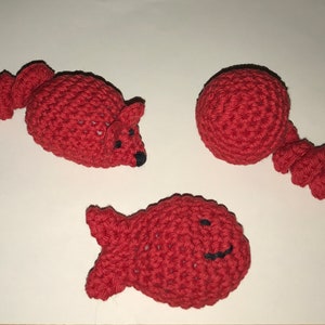 May include: Three handmade crochet toys in red: a mouse with black eyes and nose, a ball with a spiral tail, and a fish with a smiling face. These soft toys are likely designed for pets.