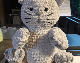 Puede incluir: Un peluche de gato hecho a mano, de ganchillo gris, con bigotes negros, ojos negros y detalles grises en las patas y las orejas. El gato está sentado sobre una superficie de madera. El juguete es suave y tierno.