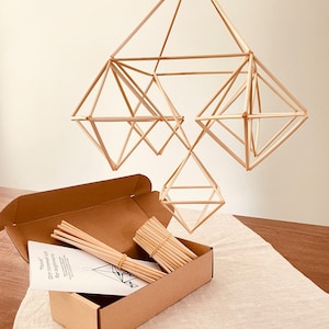 May include: A DIY geometric mobile kit with instructions and natural colored bamboo straws in a brown cardboard box. The mobile is made of three geometric shapes, a large diamond, a smaller diamond, and a cube.