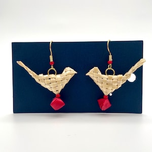 May include: A pair of handcrafted bird-shaped earrings. The earrings are woven from light tan material, with red accents and gold-colored hooks. The birds are perched on a dark blue background.