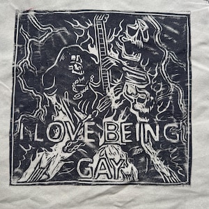 May include: Black and white graphic print on a light grey fabric. The print features a skeleton holding a guitar with the words "I LOVE BEING GAY" below.