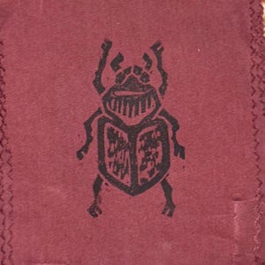 May include: A deep maroon fabric patch featuring a black, stylized block-print illustration of a beetle. The design displays a textured, ink-stamped appearance with visible jagged edges and a zigzag stitched border along the left and right sides.
