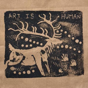 May include: A tan fabric surface featuring a black block print design of a deer with large antlers. The text ART IS HUMAN is printed at the top. The design includes circular dots, vertical line patterns, and three handprint silhouettes.