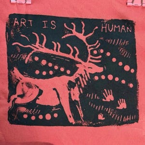 May include: A coral textile with a dark green print of a deer, dots, and handprints. The text "ART IS HUMAN" is above the deer. The design has a rustic, artistic aesthetic, reminiscent of cave paintings.