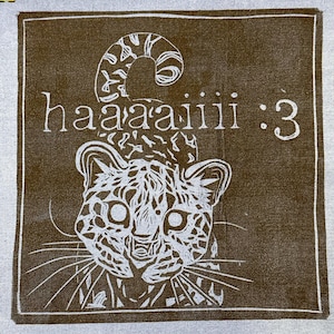 May include: A square fabric patch featuring a brown background with a white line-art illustration of a leopard. The text "haaaaiii :3" is printed in white above the leopard's head. The design is enclosed within a thin white rectangular border.