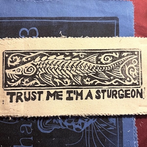 May include: A rectangular off-white fabric patch featuring a black ink block print of a sturgeon fish surrounded by decorative scrollwork. Below the fish, the text reads, "TRUST ME I'M A STURGEON" in a bold, blocky, hand-stamped style font.