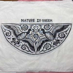 Puede incluir: Tela blanca con una impresión negra de dos pájaros enfrentados, con el texto "NATURE IS QUEER" encima. Los pájaros están rodeados de remolinos decorativos y formas de corazón.