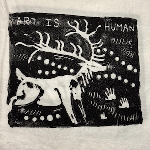 Op de afbeelding: Een zwart-witte blokdruk op stof met een gestileerd hert met groot gewei. De tekst ART IS HUMAN staat bovenaan gedrukt. Het ontwerp bevat cirkelvormige stippen en handafdrukken die rondom het hert op een donkere achtergrond zijn geplaatst.