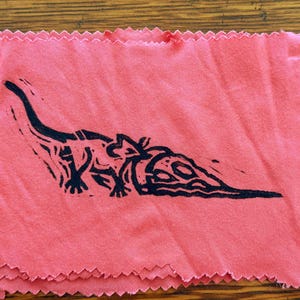 Pode incluir: Um pedaço de tecido rosa com uma ilustração preta de um lagarto ou criatura semelhante. O tecido tem uma borda bruta e inacabada. Um clip de papel prateado está preso ao tecido.