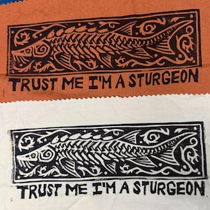 Peut inclure: Deux rectangles de tissu, un orange et un blanc, présentent un motif d'esturgeon noir et blanc avec le texte "TRUST ME I'M A STURGEON". L'illustration de l'esturgeon est encadrée d'une bordure décorative.