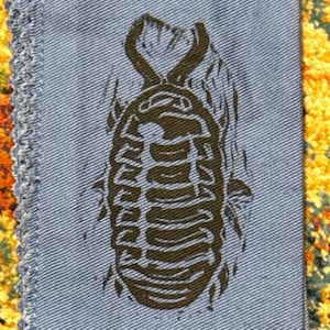 Puede incluir: Primer plano de tela vaquera azul claro con un diseño de insecto marrón oscuro. El insecto es una ilustración estilizada de una cochinilla. El borde de la tela tiene un ribete decorativo con flecos.