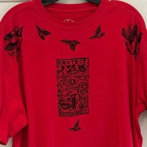 May include: A red long-sleeved t-shirt with black bird illustrations and the words "PROTECT BIODIVERSITY". The design is in a rectangular shape. The shirt has a crew neck and is made of a soft material.