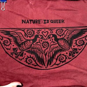 Puede incluir: Tela granate con el texto negro "NATURE IS QUEER". Debajo, un gráfico negro de dos pájaros enfrentados, con un corazón entre ellos. Los pájaros están rodeados de remolinos y hojas decorativas.