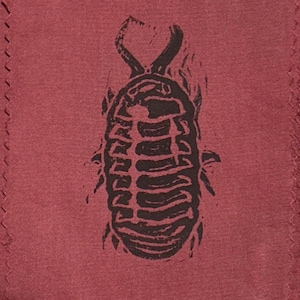 May include: A dark red fabric featuring a black block-print style graphic of a pill bug or woodlouse. The insect illustration is centered on the textile, displaying a segmented oval body with small legs and two antennae at the top of the design.