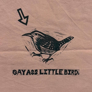 May include: A linocut print of a bird with an arrow pointing towards it. The bird is black and white with detailed feathers. Below the bird is the text "GAY ASS LITTLE BIRD" in a bold font. The background is a light brown colour.