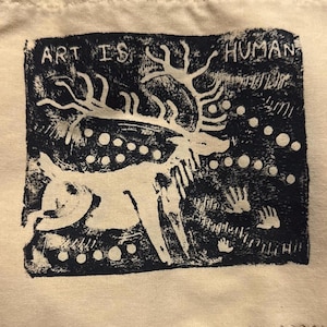 May include: A cream-colored textile featuring a black and white print. The print depicts a deer with large antlers, surrounded by dots and handprints. The text "ART IS HUMAN" is printed above the deer.