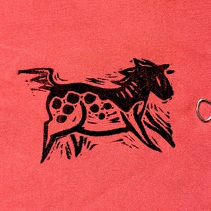 May include: A black and white linocut print of a horse with a spotted body and flowing mane, set against a coral-colored background. The horse is depicted in motion, with its legs extended as if running.